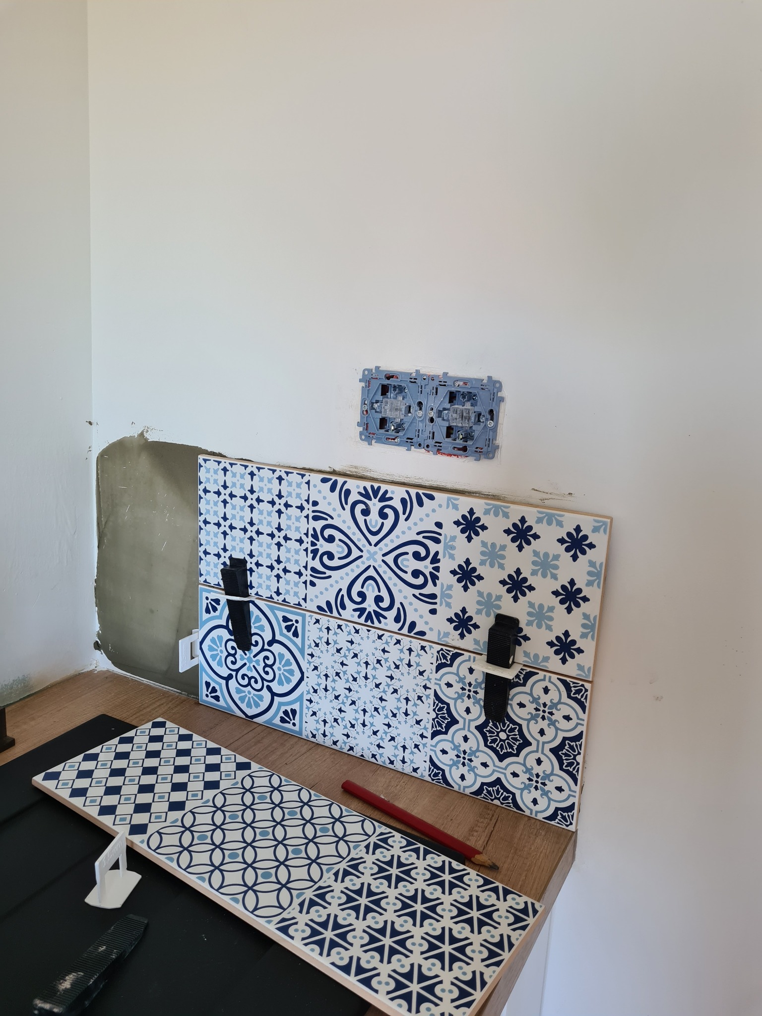 Fragment ściany z częściowo ułożonymi płytkami dekoracyjnymi w odcieniach błękitu i granatu, widoczne narzędzia glazurnicze, puszka elektryczna nad płytkami.