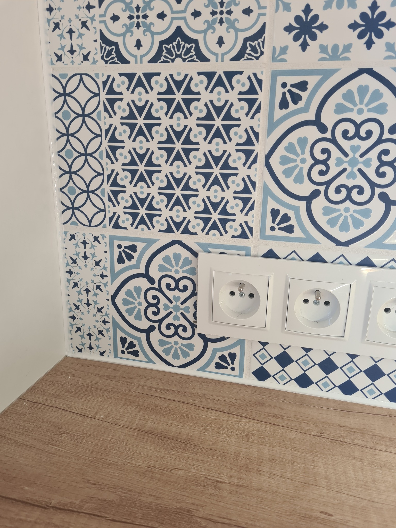 Ściana wyłożona dekoracyjnymi, niebiesko-białymi płytkami ceramicznymi z motywami geometrycznymi i kwiatowymi, z trzema gniazdkami elektrycznymi i fragmentem drewnianego blatu.