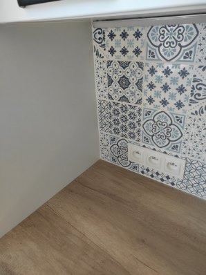 Fragment kuchni po remoncie: blat drewnopodobny, ściana z dekoracyjnych płytek w odcieniach błękitu i szarości oraz trzy gniazdka elektryczne w białej ramce.