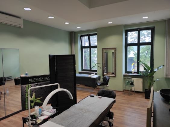 Jasne wnętrze gabinetu kosmetycznego z dwoma łóżkami do zabiegów, parawanem, lustrem w złotej ramie i dużą rośliną doniczkową przy oknie.