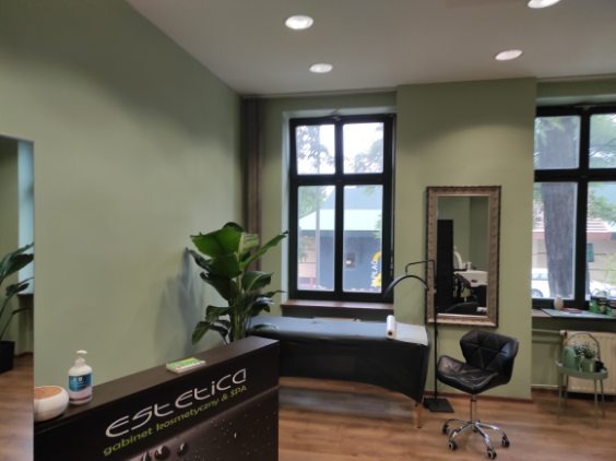 Wnętrze gabinetu kosmetycznego z zielonymi ścianami, widok na kozetkę, czarne krzesło na kółkach i rośliny doniczkowe, w tle okna.