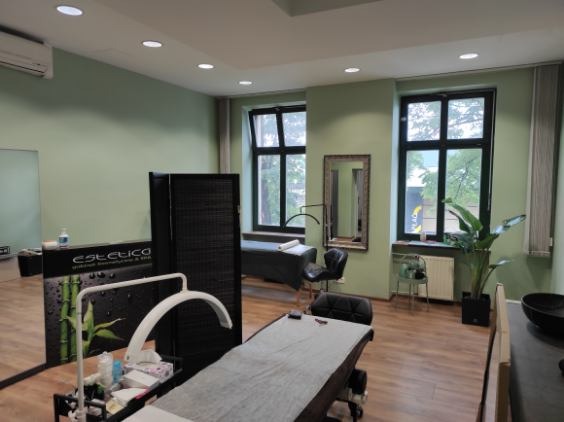 Wnętrze gabinetu kosmetycznego z dwoma stanowiskami do zabiegów, czarnym parawanem, lustrem w drewnianej ramie i dużymi oknami z widokiem na zieleń.