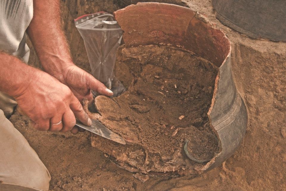 Ostrożne wydobywanie zabytkowych fragmentów naczynia z ziemi przy użyciu szpachelki, obok woreczek na znaleziska. Prace archeologiczne w toku.