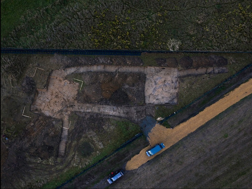 Zdjęcie z drona przedstawia wykop pod fundamenty domu w kształcie litery T, z dwoma zaparkowanymi samochodami w pobliżu i polami w tle.