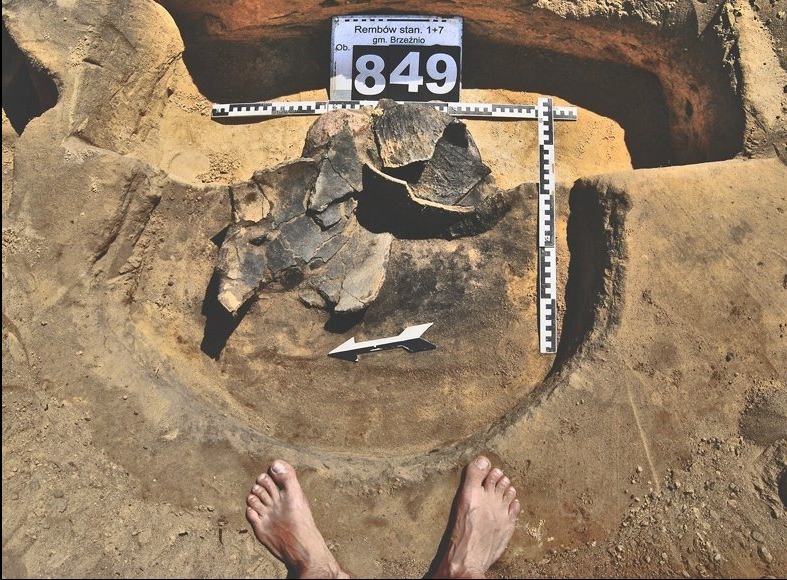 Stanowisko archeologiczne: wykop z ceramiką, miarką i tabliczką identyfikacyjną. W kadrze stopy osoby. Wykop w ziemi o okrągłym kształcie.