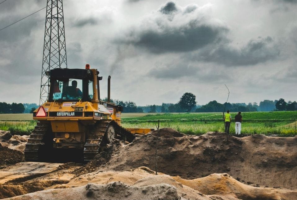 Żółty spychacz Caterpillar na placu budowy, dwóch mężczyzn w tle, pochmurne niebo. Widoczna wieża wysokiego napięcia w oddali.