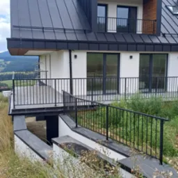 Nowoczesny dom w Łętowicach z czarną metalową balustradą na balkonie i przy zejściu do piwnicy, otoczony wysoką trawą.