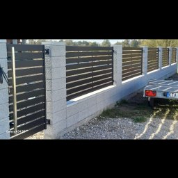 FHU SPAW Smoszna Janusz - Nowoczesne ogrodzenie z poziomych, ciemnych paneli zamontowanych między szarymi słupkami z bloczków betonowych, widoczny fragment przyczepki samochodowej w tle.