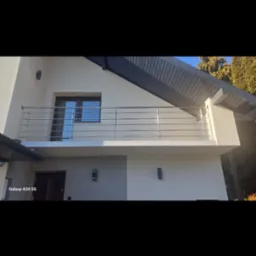 Balkon z nowoczesną, poziomą balustradą ze stali nierdzewnej, zamontowaną na jasnym budynku z ciemnym dachem i oknem.