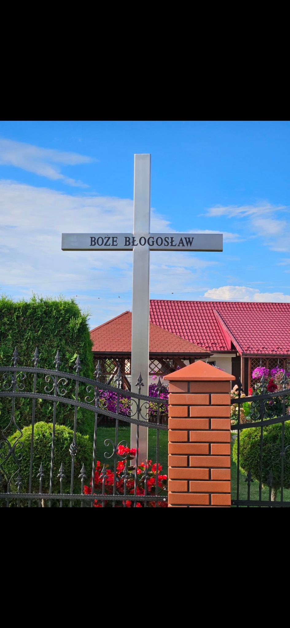Metalowy krzyż z napisem 'Boże Błogosław' na tle błękitnego nieba, widoczny zza kutego ogrodzenia w Łętowicach, z elementami muru z cegły klinkierowej i zieleni w tle.