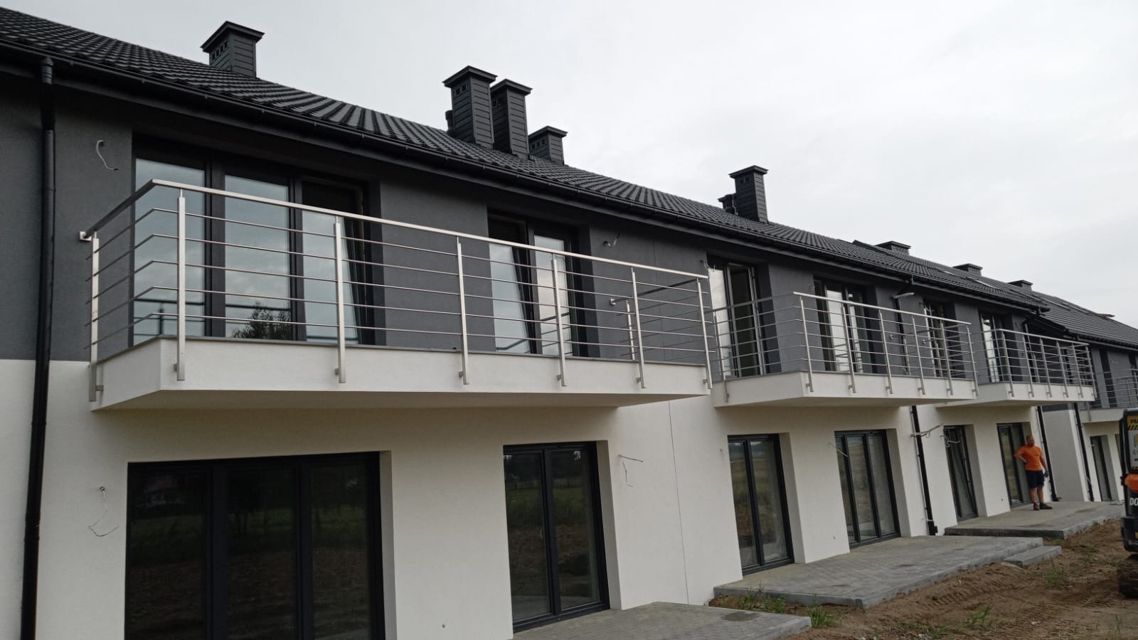 Nowoczesne balkony szeregowca z prostymi, metalowymi barierkami, widok z zewnątrz na szary budynek z czarnym dachem.