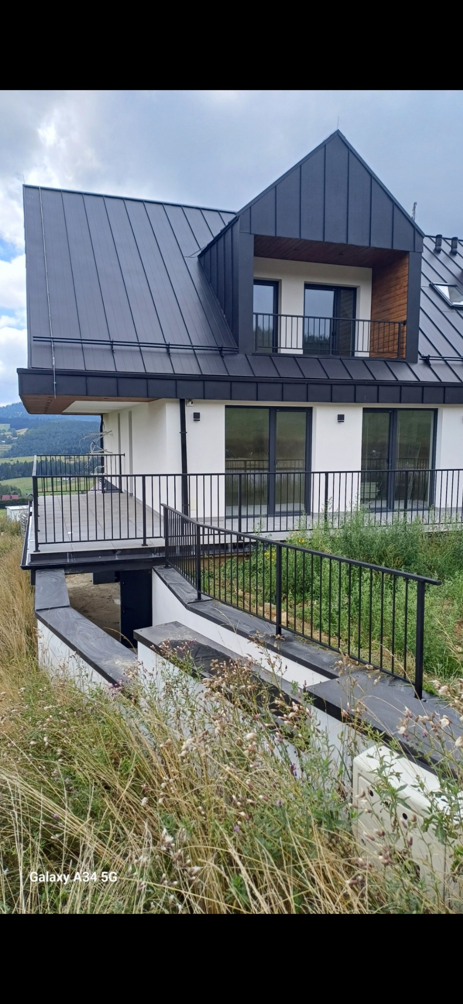 Nowoczesny dom w Łętowicach z czarną metalową balustradą na balkonie i przy zejściu do piwnicy, otoczony wysoką trawą.