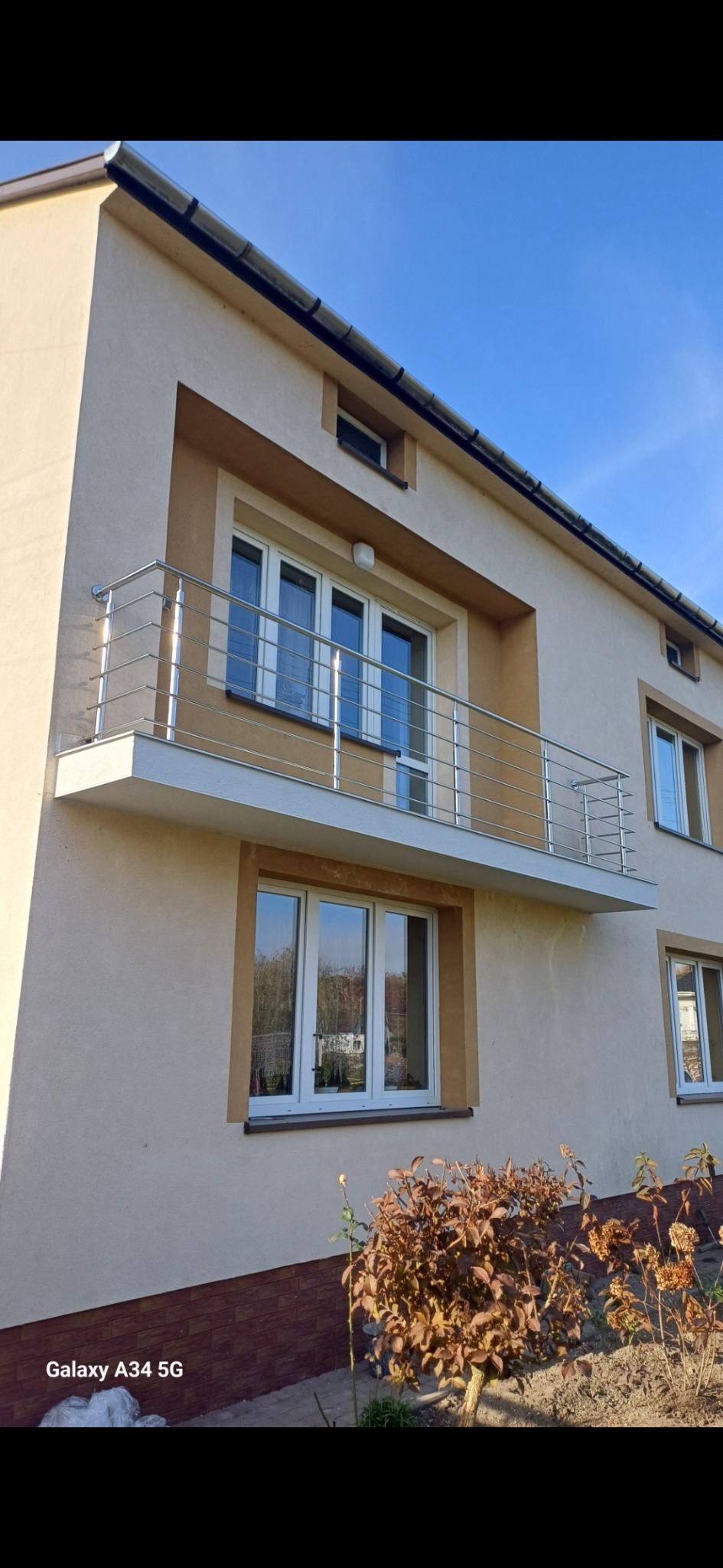 Dwupiętrowy dom z beżową elewacją i balkonem z metalową, minimalistyczną barierką, widok z dołu na błękitne niebo.