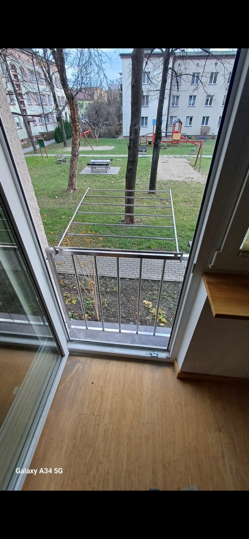 Widok z balkonu z niską, nierdzewną barierką, na plac zabaw z piaskownicą i stolikiem piknikowym, przez otwarte okno balkonowe.