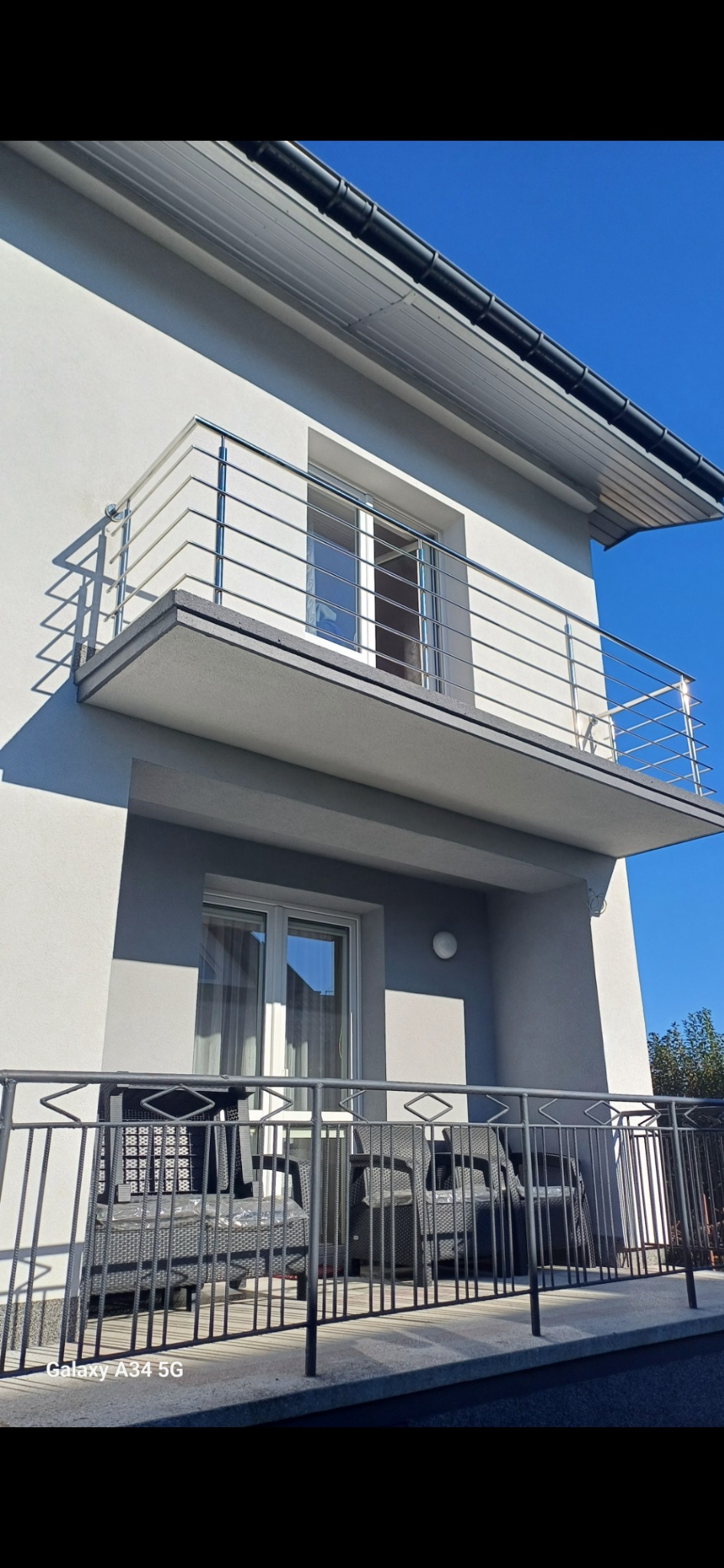 Nowoczesny dom z balkonem i metalową balustradą, widok z dołu na tle błękitnego nieba.