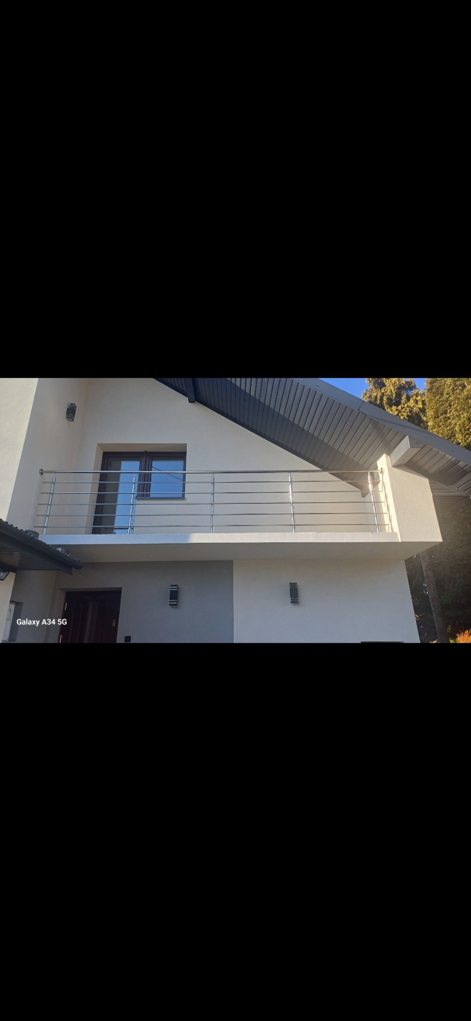 Balkon z nowoczesną, poziomą balustradą ze stali nierdzewnej, zamontowaną na jasnym budynku z ciemnym dachem i oknem.