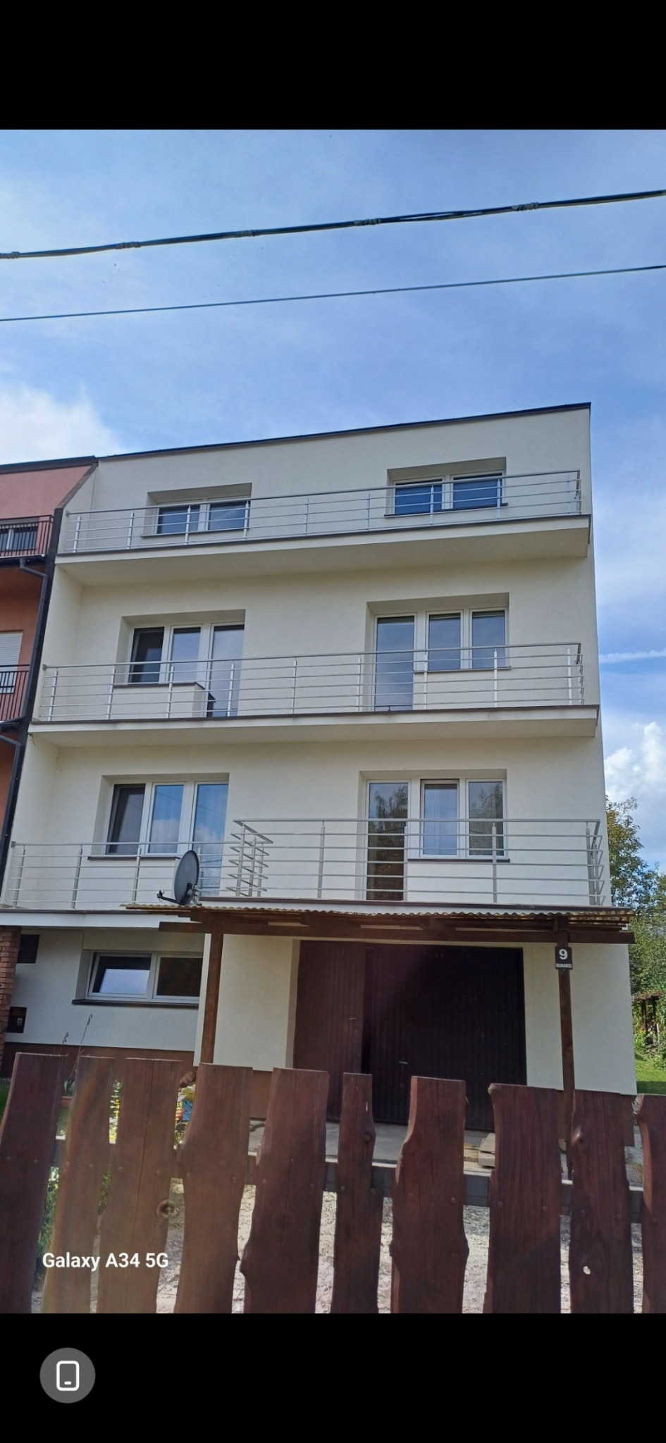 Trzykondygnacyjny dom z jasną elewacją i balkonami z nierdzewnymi balustradami. Widok zza drewnianego ogrodzenia.