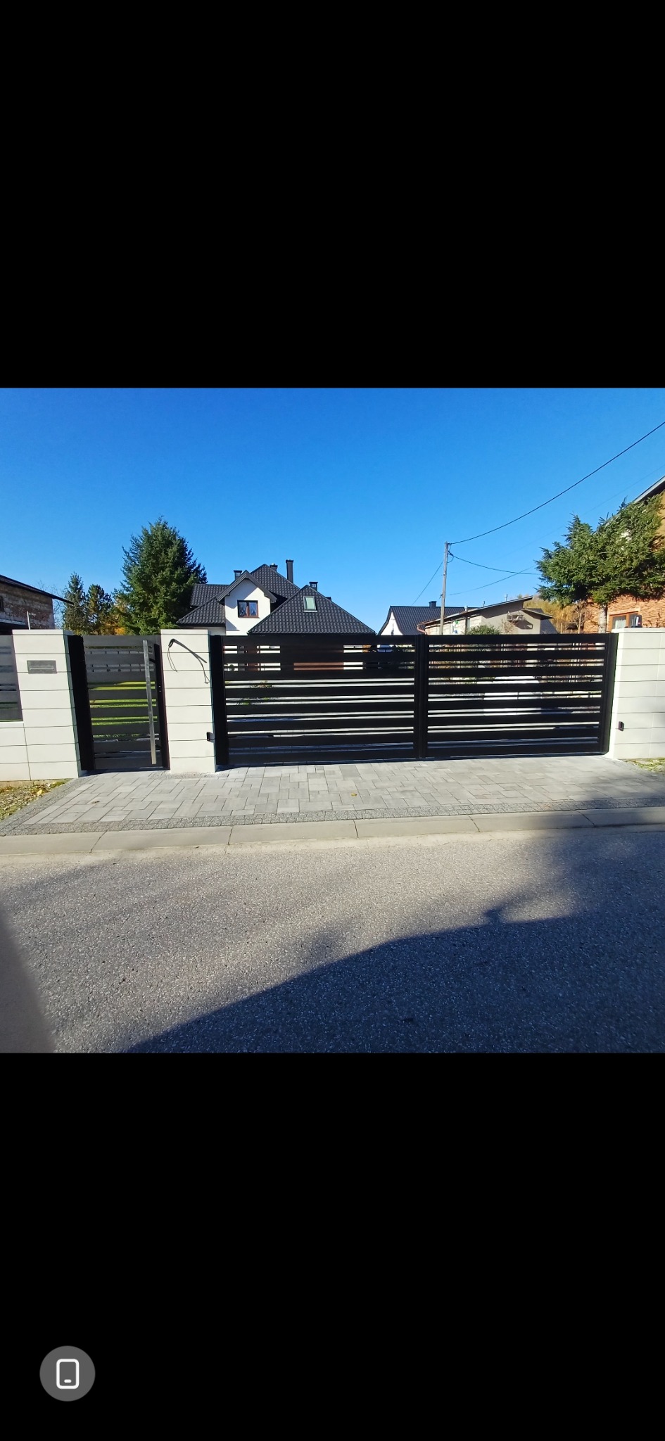 Nowoczesna brama wjazdowa z poziomymi, czarnymi panelami na tle błękitnego nieba i domu. Brama zamontowana na betonowych słupkach, z kostką brukową przed nią.