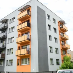 Wielopiętrowy budynek mieszkalny z balkonami o pomarańczowych i szarych balustradach, widok z dołu, zadaszenia nad balkonami.