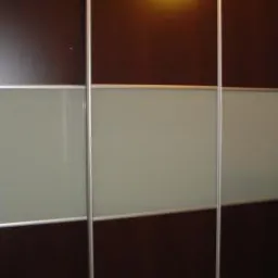 Trzydrzwiowa szafa przesuwna z ciemnobrązowymi panelami i mlecznymi szybami, obramowana srebrnymi profilami.