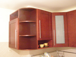 Ciemnobrązowa, narożna szafka kuchenna na wymiar z półkami i przeszklonym frontem, widoczne filiżanki na półce, oświetlenie sufitowe.