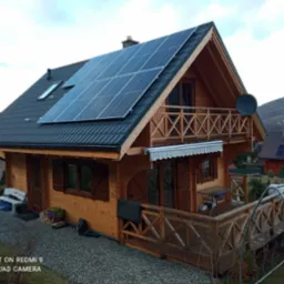 Drewniany dom z ciemnym dachem i panelami słonecznymi na tle górskim, z balkonem i satelitą.