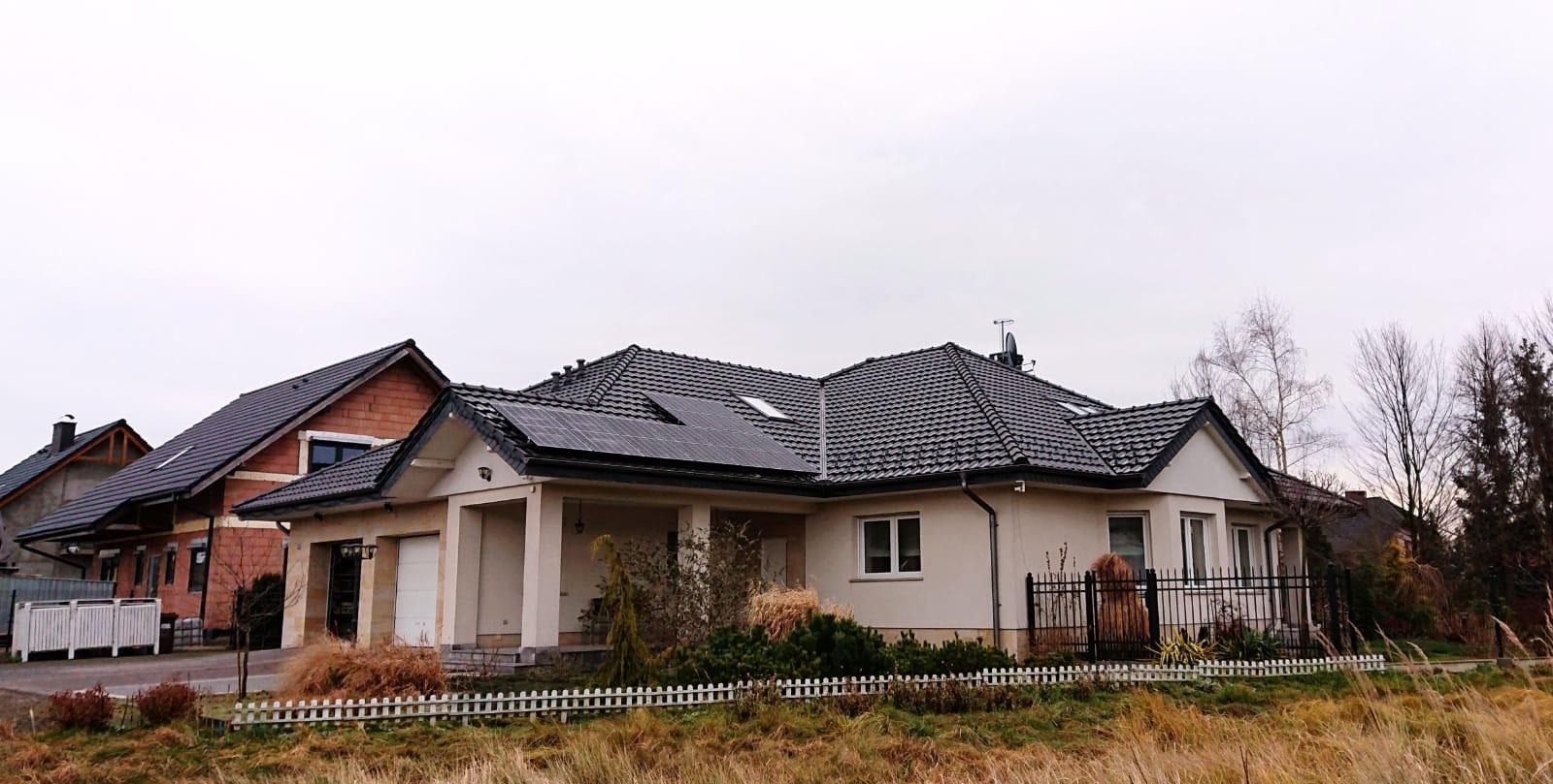 Jednorodzinny dom z ciemną dachówką i panelami słonecznymi na dachu, widoczny z perspektywy trawnika przed posesją w pochmurny dzień.