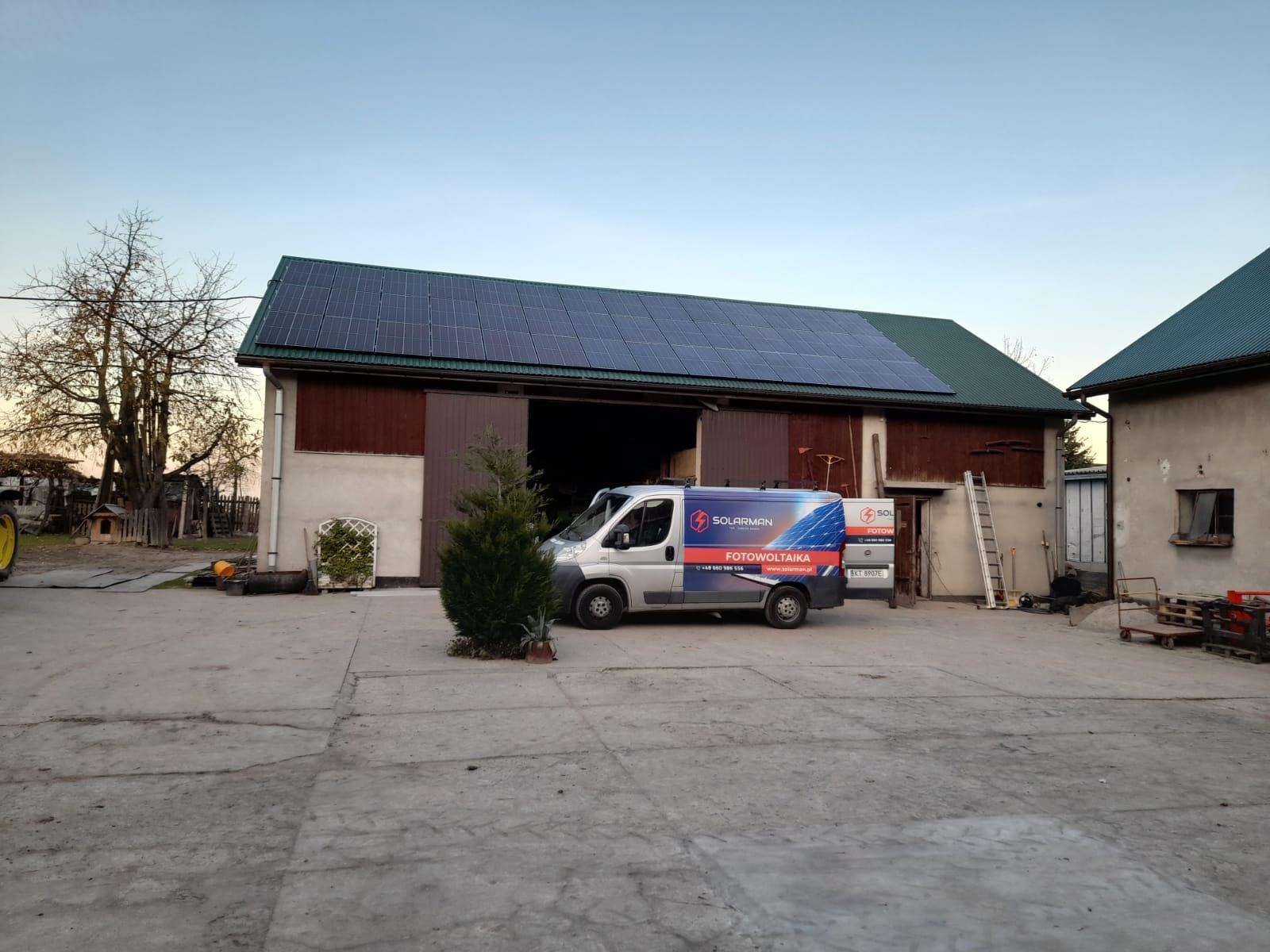 Instalacja paneli słonecznych na dachu budynku gospodarczego; na pierwszym planie samochód dostawczy z logo firmy instalującej.
