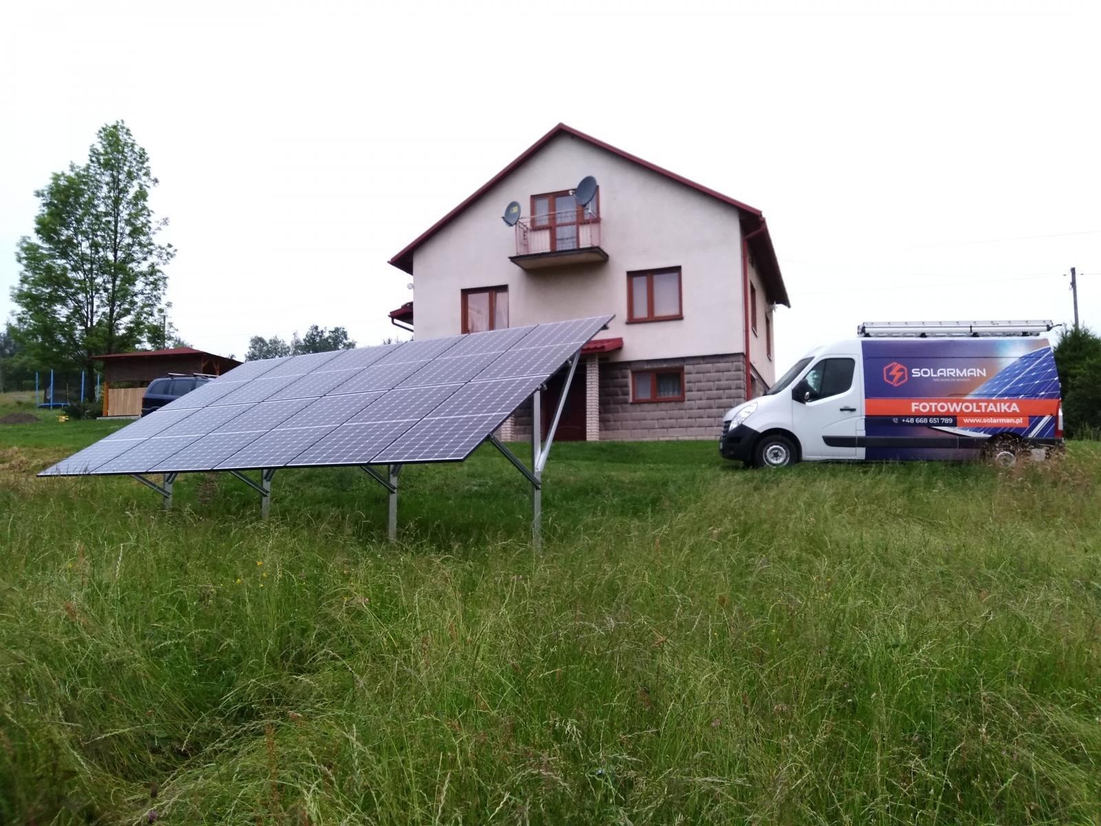 Panel słoneczny na metalowej konstrukcji przed domem jednorodzinnym, obok zaparkowany biały bus z logo firmy instalującej panele.