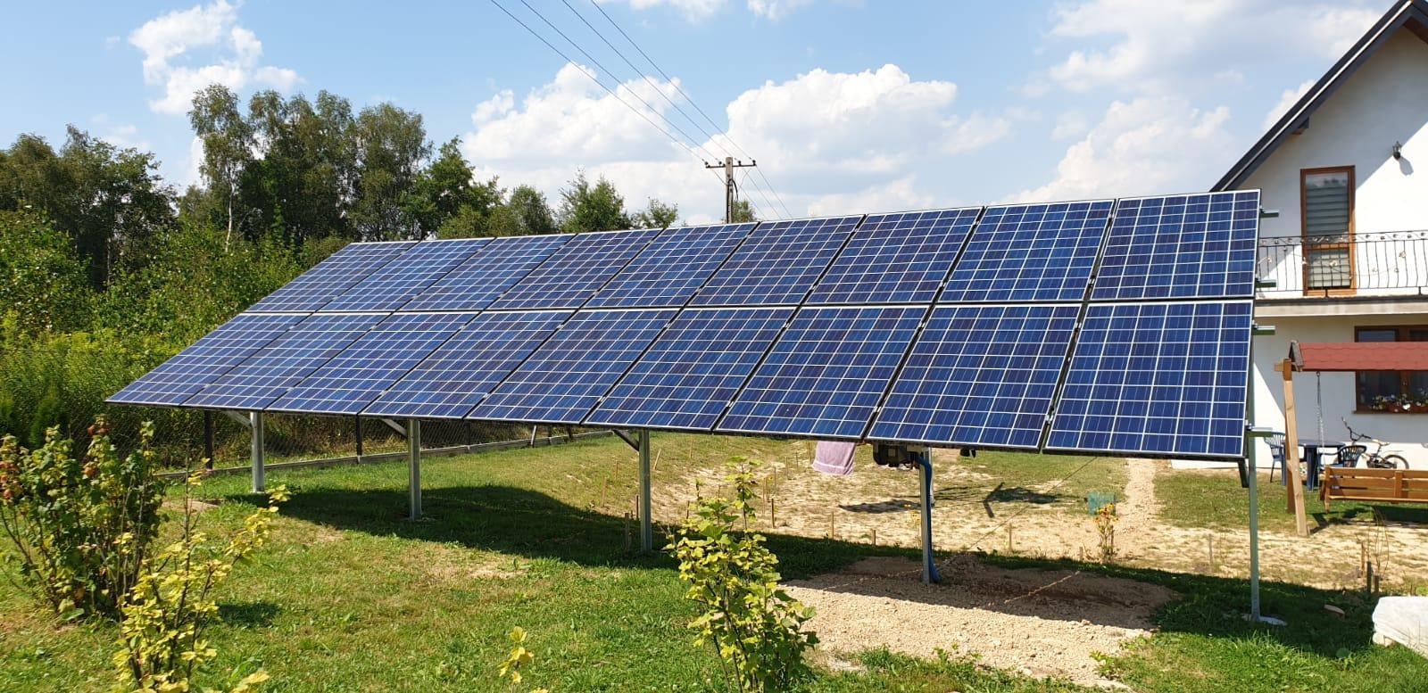 Instalacja fotowoltaiczna na stelażu przed domem jednorodzinnym, widoczne panele w rzędach, trawa i drzewa w tle.