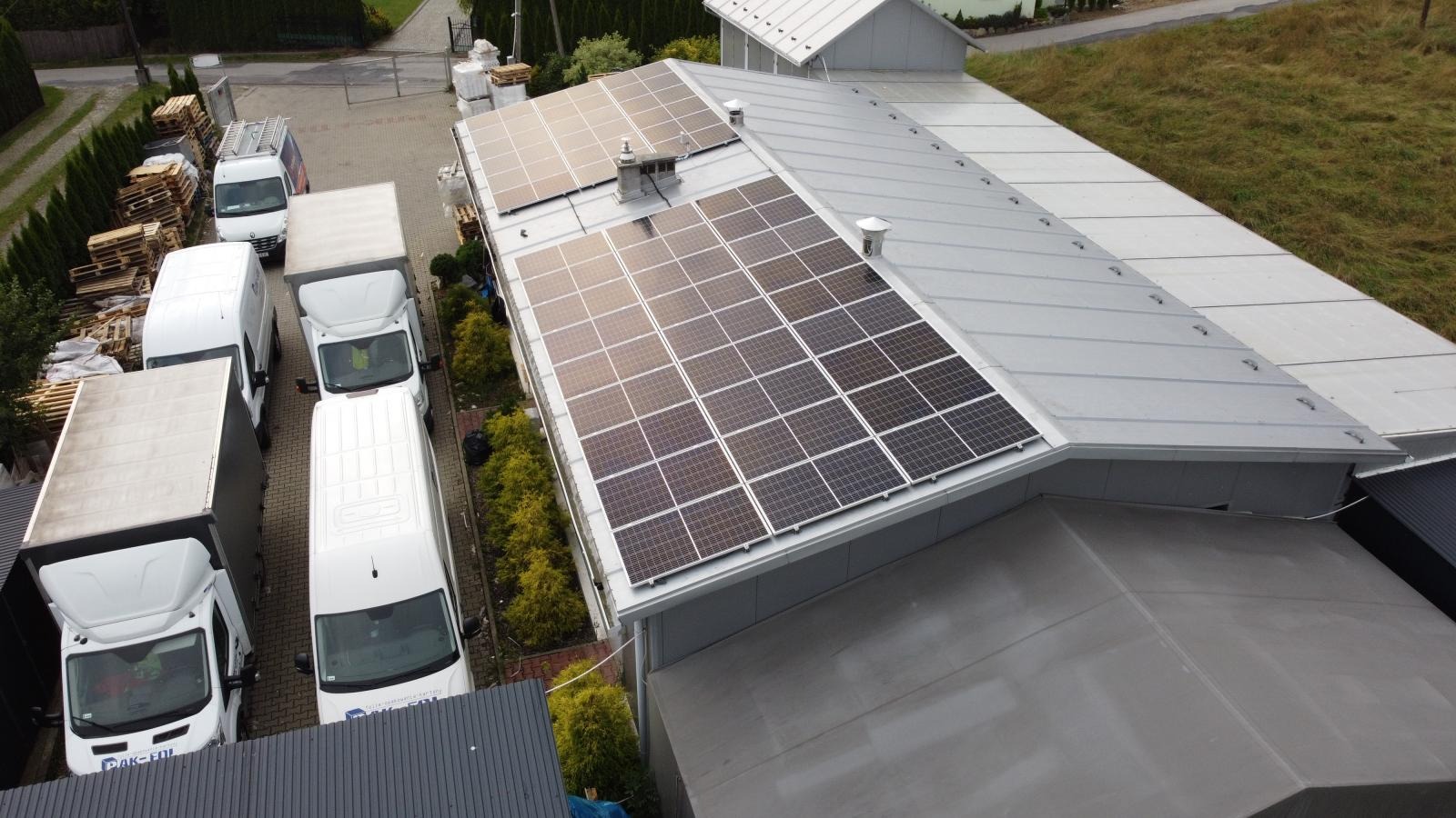 Widok z góry na zainstalowane panele słoneczne na szarym dachu budynku przemysłowego, z zaparkowanymi białymi samochodami dostawczymi w tle.
