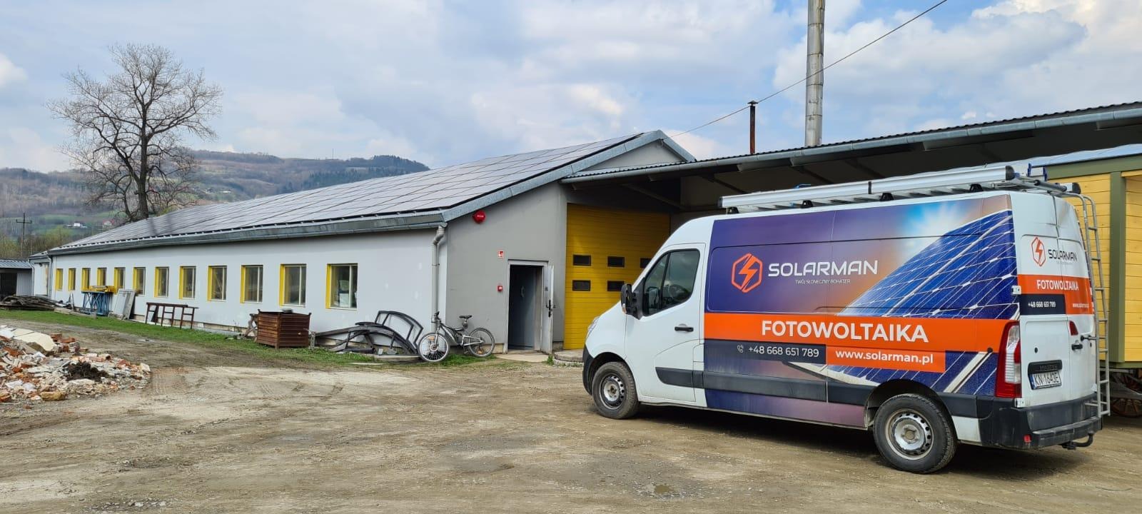 Budynek gospodarczy z panelami słonecznymi na dachu, obok zaparkowany biały samochód dostawczy z reklamą firmy fotowoltaicznej Solarman, widoczne góry w tle.