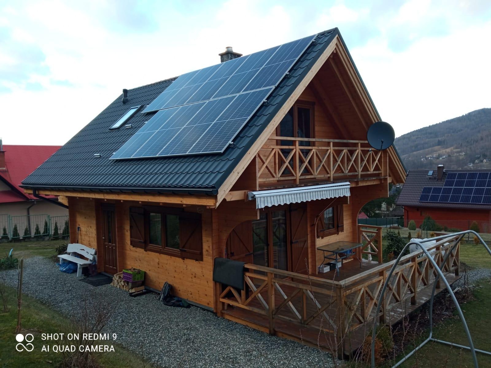 Drewniany dom z ciemnym dachem i panelami słonecznymi na tle górskim, z balkonem i satelitą.