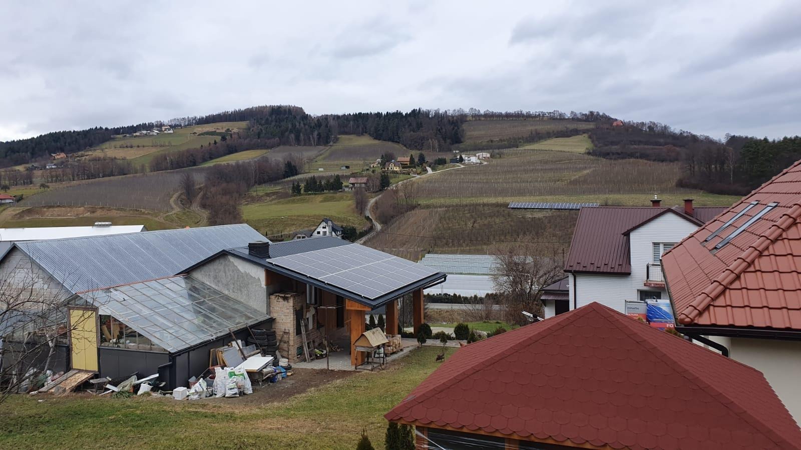 Widok z góry na wiejski krajobraz z panelami słonecznymi na dachu domu, w tle wzgórza z winnicami i pochmurne niebo.