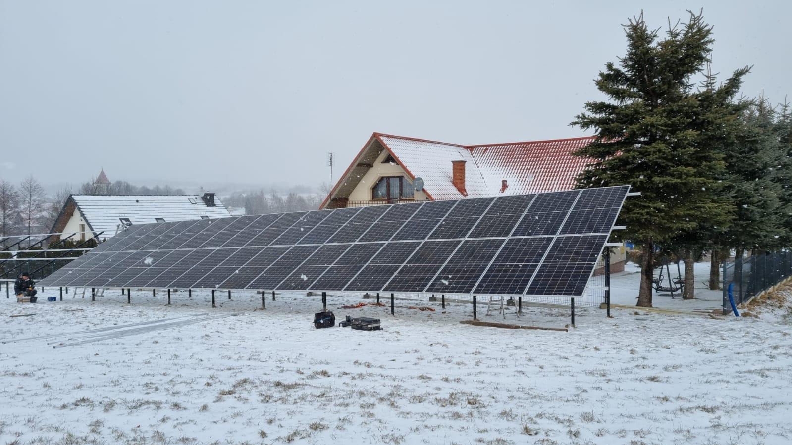 Panele słoneczne ustawione na śniegu przed domem z czerwoną dachówką, widoczna instalacja wsporcza i skrzynki narzędziowe.