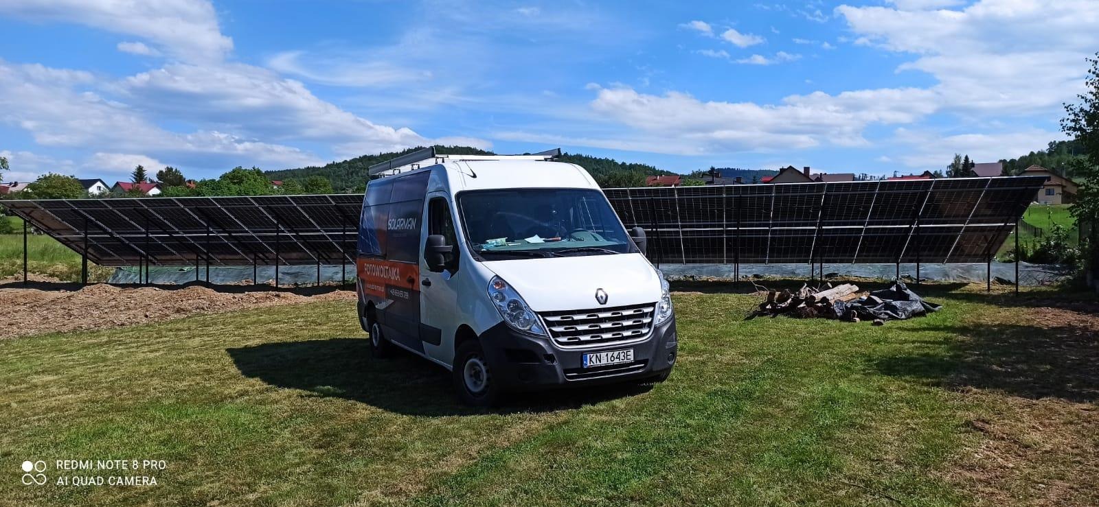 Biały samochód dostawczy z logo firmy na tle zainstalowanych paneli słonecznych na trawiastym terenie w słoneczny dzień.