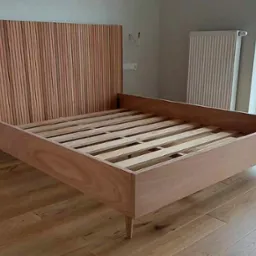 Drewniane łóżko z listwowym wezgłowiem w surowym wykończeniu, widoczne elementy konstrukcyjne i stelaż, ustawione na parkiecie w jasnym wnętrzu z oknem.