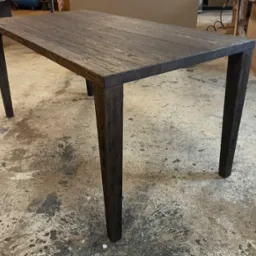 Stół z ciemnego drewna o wyraźnym usłojeniu, proste nogi, surowa podłoga warsztatowa w tle.