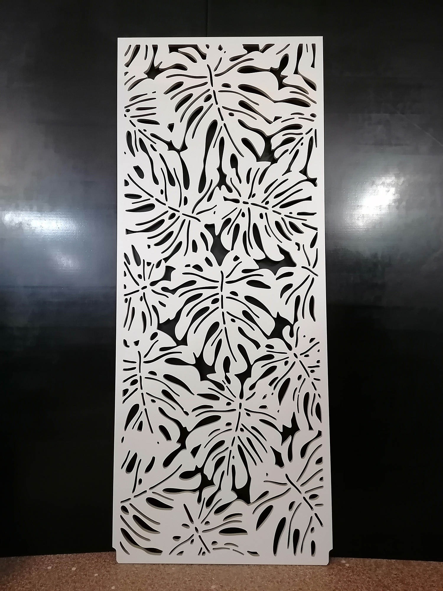 Biały, ażurowy panel dekoracyjny z motywem liści monstery na ciemnym tle, stojący na podłodze.