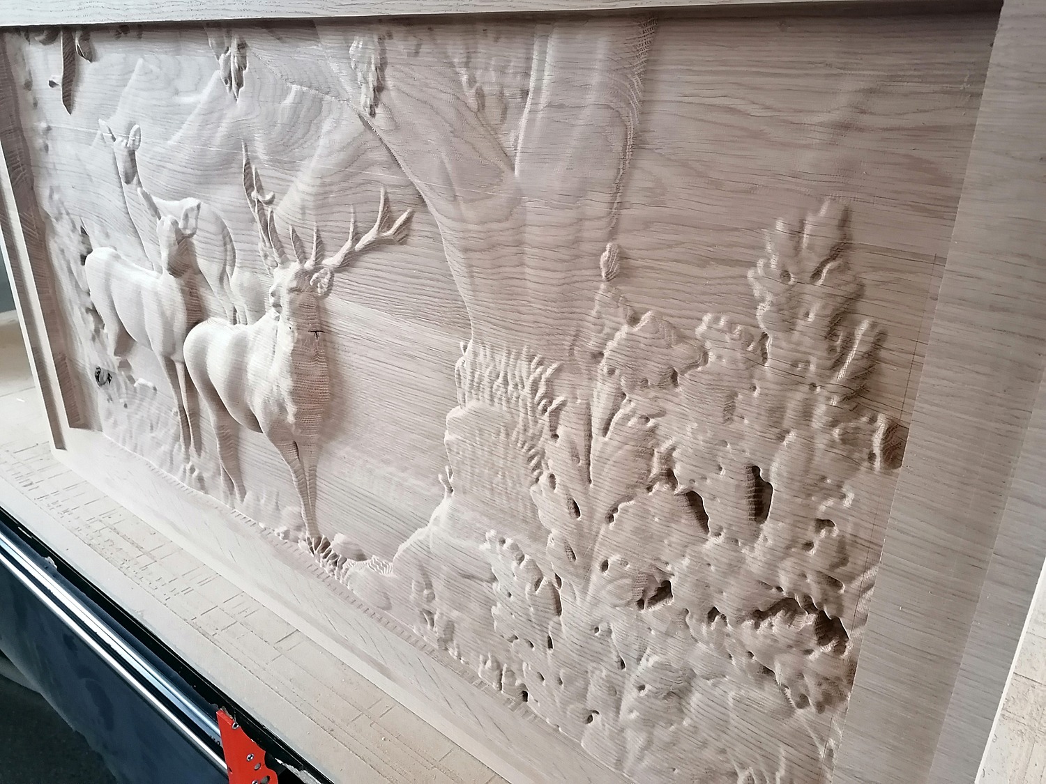 Drewniana płaskorzeźba przedstawiająca scenę z jeleniami w lesie, wykonana z jasnego drewna, widoczna pod kątem z boku, ukazująca detale rzeźbienia.