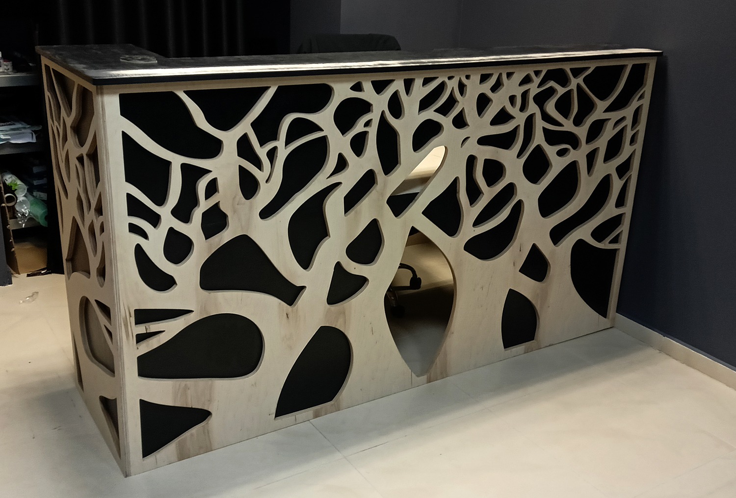 Recepcyjna lada z drewnianą obudową i ażurowym wzorem drzewa na czarnym tle, widoczna z bliska, z fragmentem biurowego wnętrza w tle.