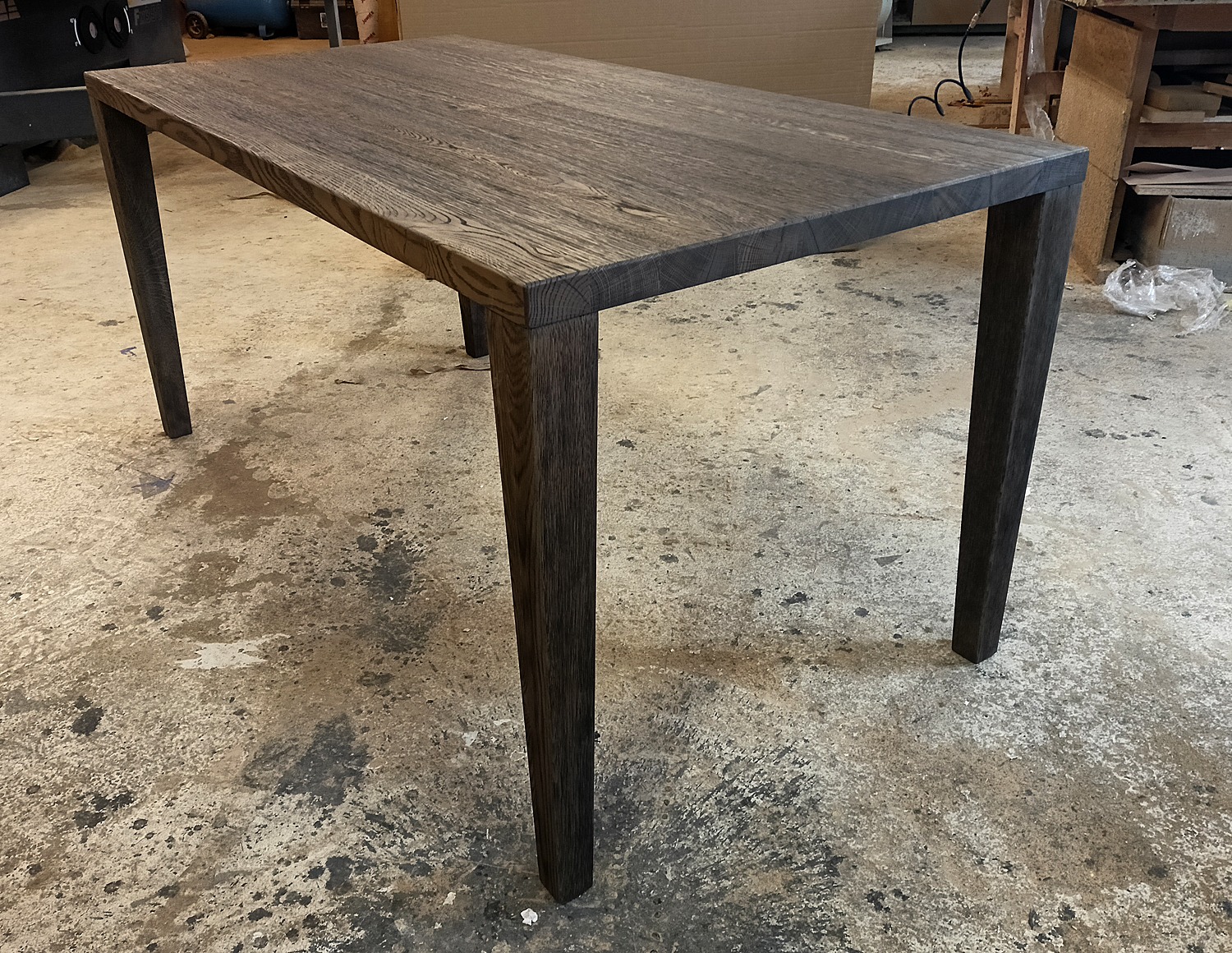 Stół z ciemnego drewna o wyraźnym usłojeniu, proste nogi, surowa podłoga warsztatowa w tle.
