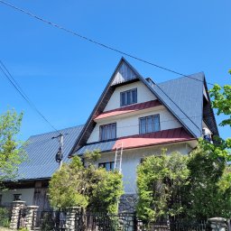 KarMal - Dwupiętrowy dom z nowym, błyszczącym, ciemnoszarym dachem z blachy, z widoczną drabiną opartą o elewację i osobą pracującą na dachu, na tle błękitnego nieba i zieleni drzew.