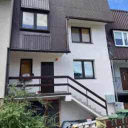 Dwupiętrowy dom z białymi ścianami i brązowym dachem z blachy trapezowej, widoczne prace na balkonie i drabina oparta o ogrodzenie, sugerujące remont elewacji.