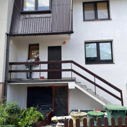 KarMal - Pracownik maluje białą elewację domu z brązowymi oknami i dachem, zabezpieczone folią drzwi wejściowe na parterze.