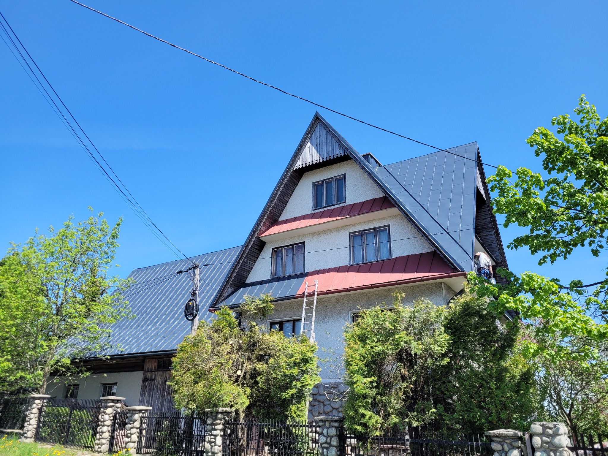 Dwupiętrowy dom z nowym, błyszczącym, ciemnoszarym dachem z blachy, z widoczną drabiną opartą o elewację i osobą pracującą na dachu, na tle błękitnego nieba i zieleni drzew.