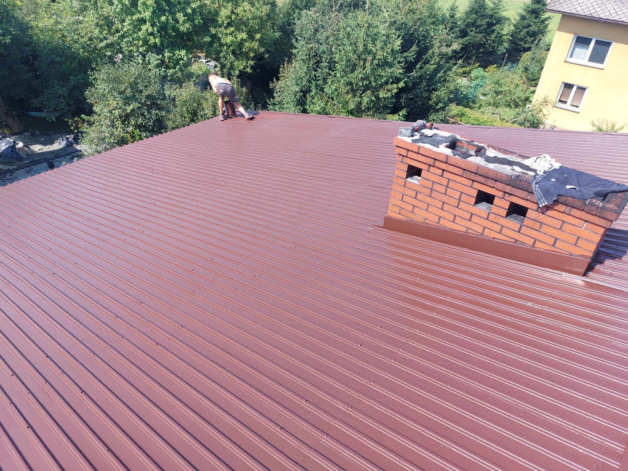 Pracownik w krótkich spodenkach maluje metalowy dach na brązowo, widoczny ceglany komin i otoczenie z zielenią oraz fragment budynku.