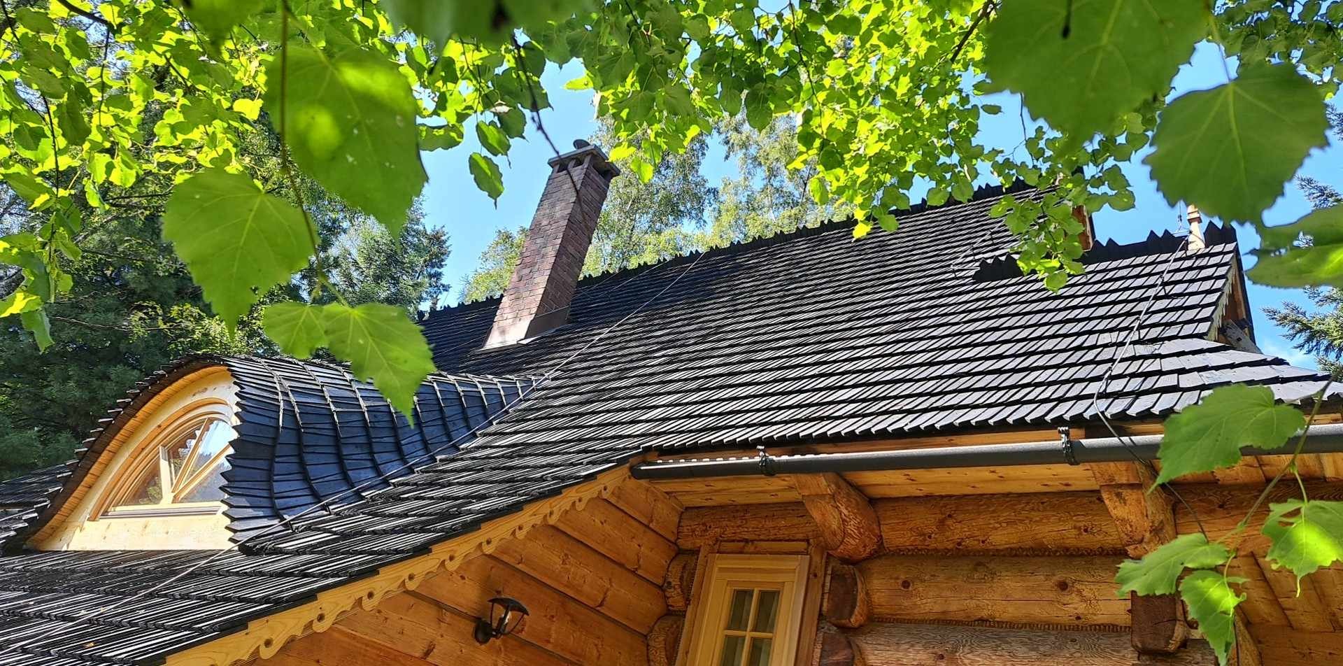 Drewniany dom z ciemnym, gontowym dachem i ceglanym kominem, widok z dołu przez liście drzew
