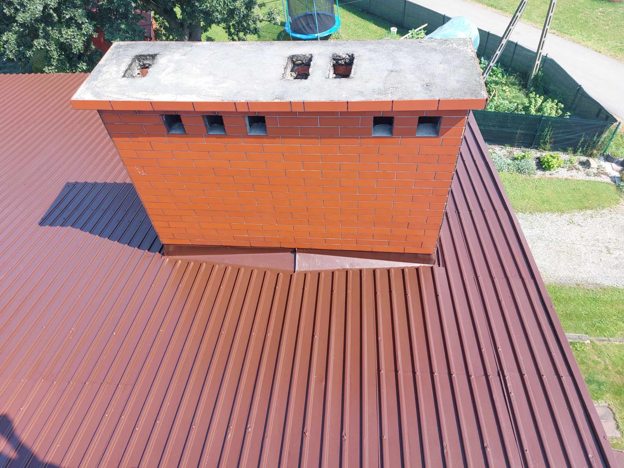 Ceglany komin z betonową czapą na brązowym, blaszanym dachu z widocznymi otworami wentylacyjnymi i uszkodzeniami na szczycie komina.