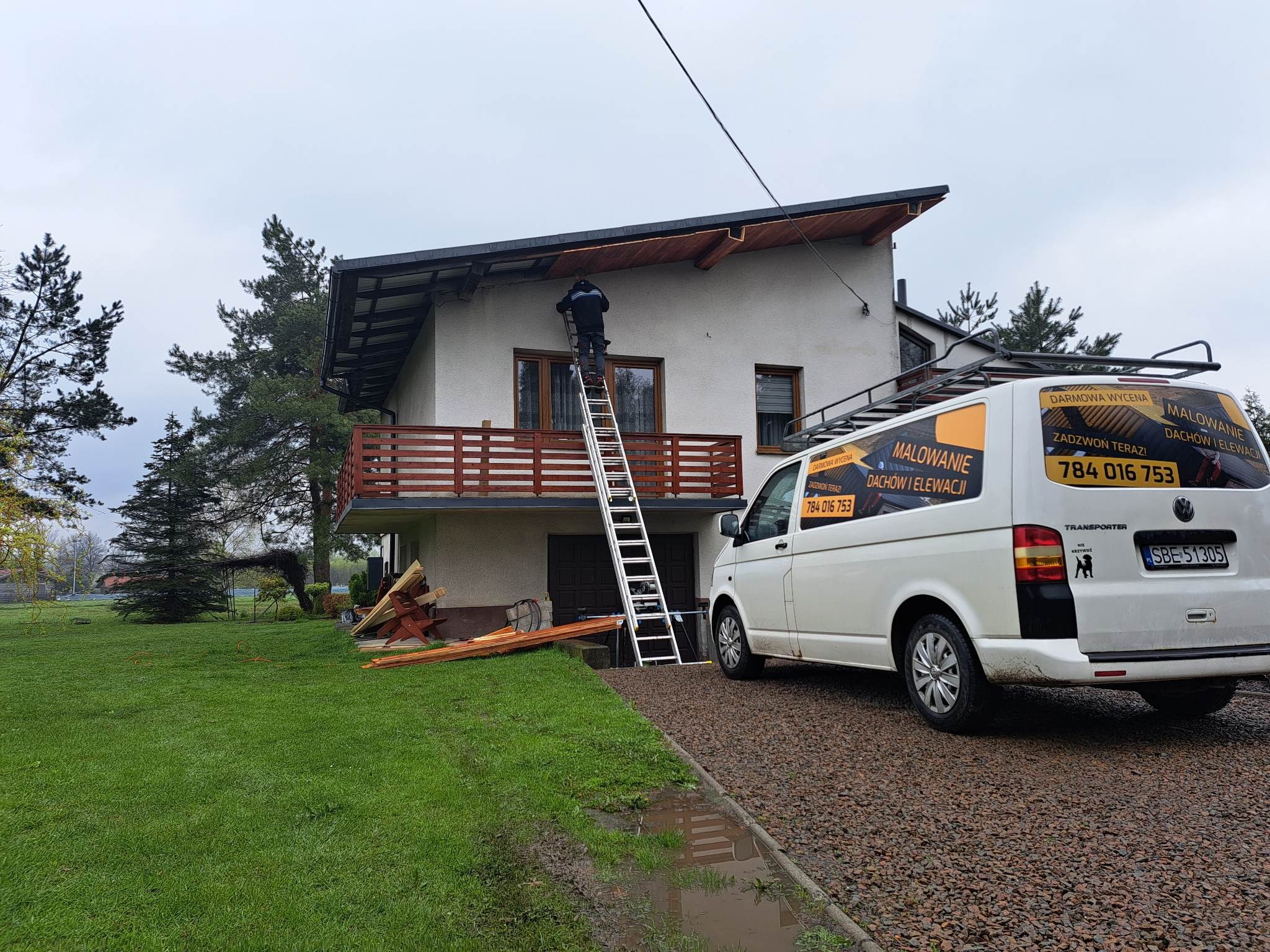 Pracownik na drabinie przy elewacji domu jednorodzinnego, obok zaparkowany bus z reklamą firmy oferującej usługi malowania dachów i elewacji, widoczne elementy drewniane przygotowane do montażu.